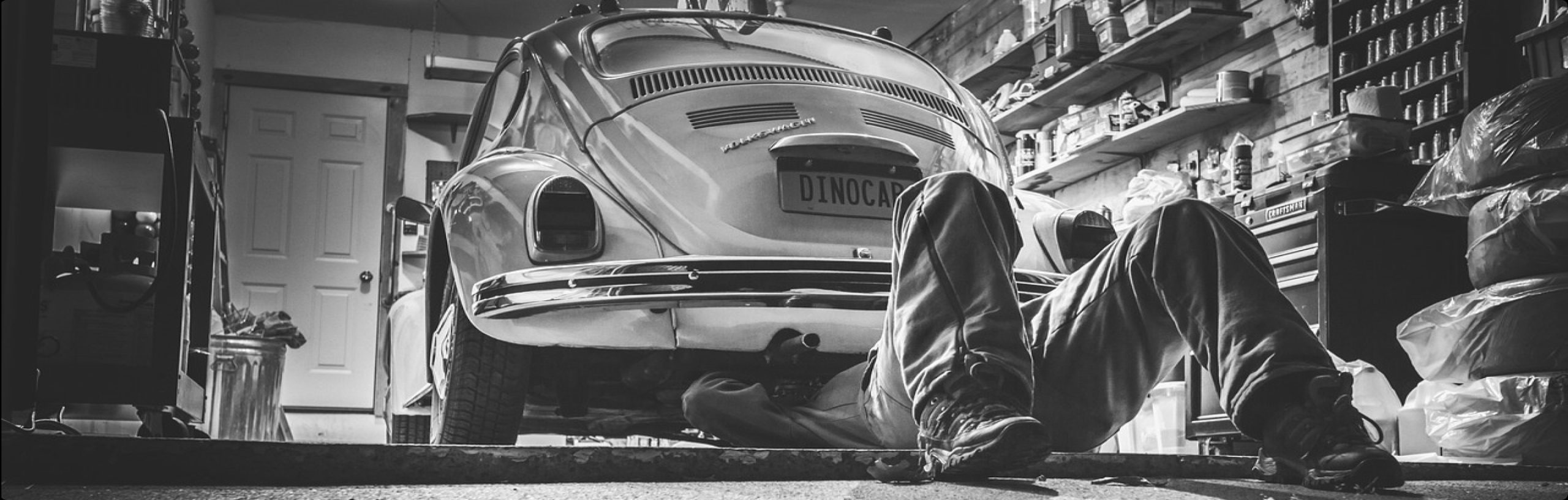 Mechanic working under a classic Volkswagen Beetle in a garage filled with tools and car parts.